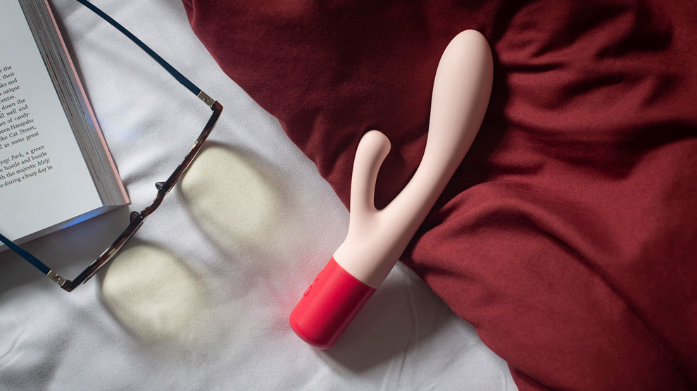 A pink durex massager toy placed on a bed beside a book and reading glasses