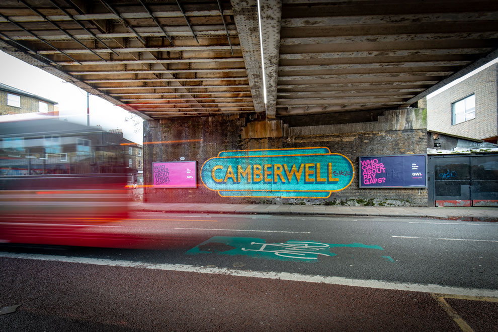 Advertising hoardings under a bridge in Camberwell, London. Bus rushing past, with an large decorative 'Camberwell' sign on the bridge support.