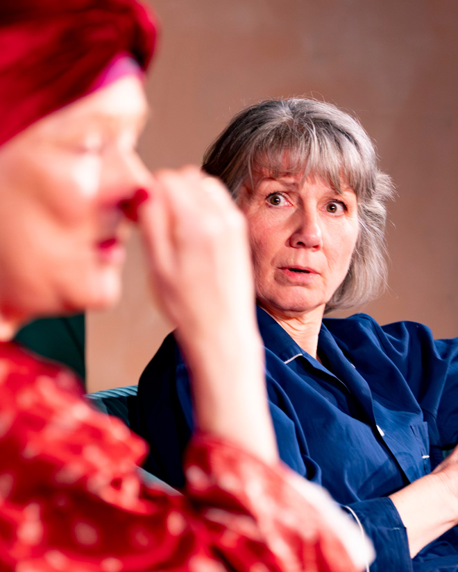 Grey-haired woman in blue looks surprised while another woman in red sits blurred in the foreground.