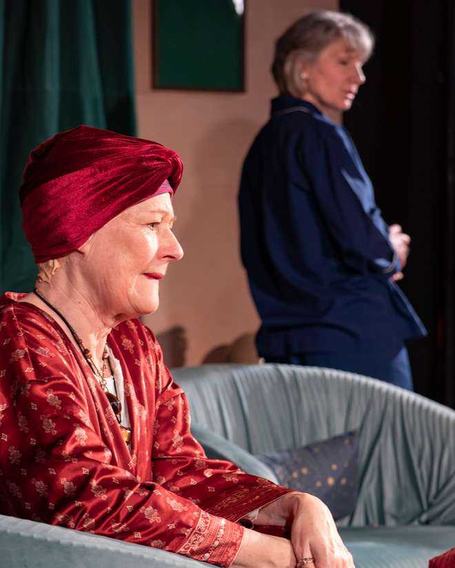 Woman in a red robe and headwrap sits on a chair while another woman stands in the background, turned away.