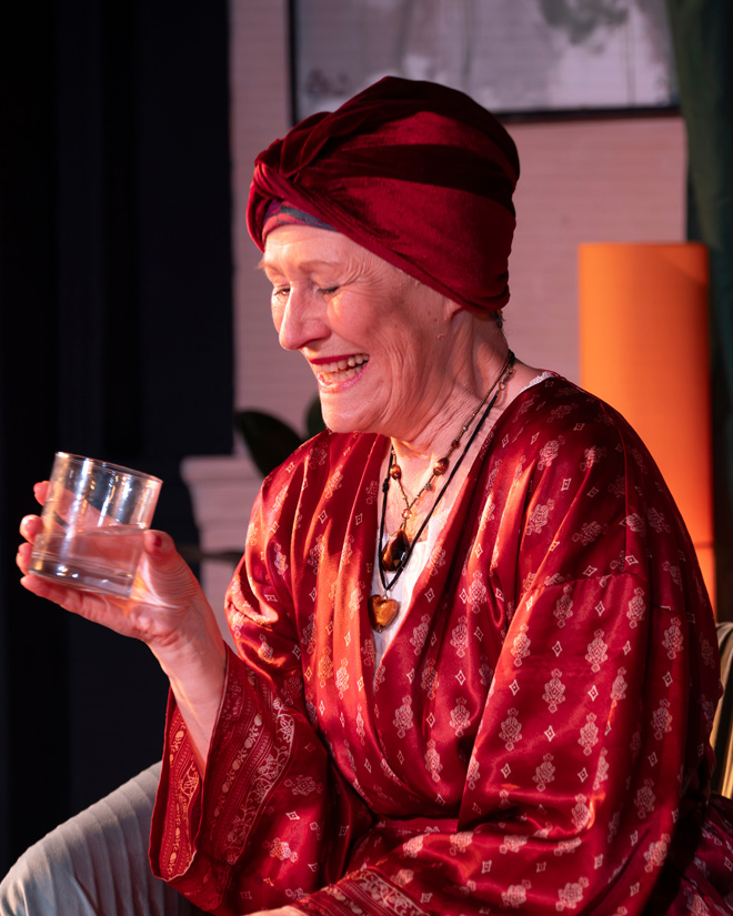 Woman in a red robe smiling and holding a glass, lit warmly against a dark background.