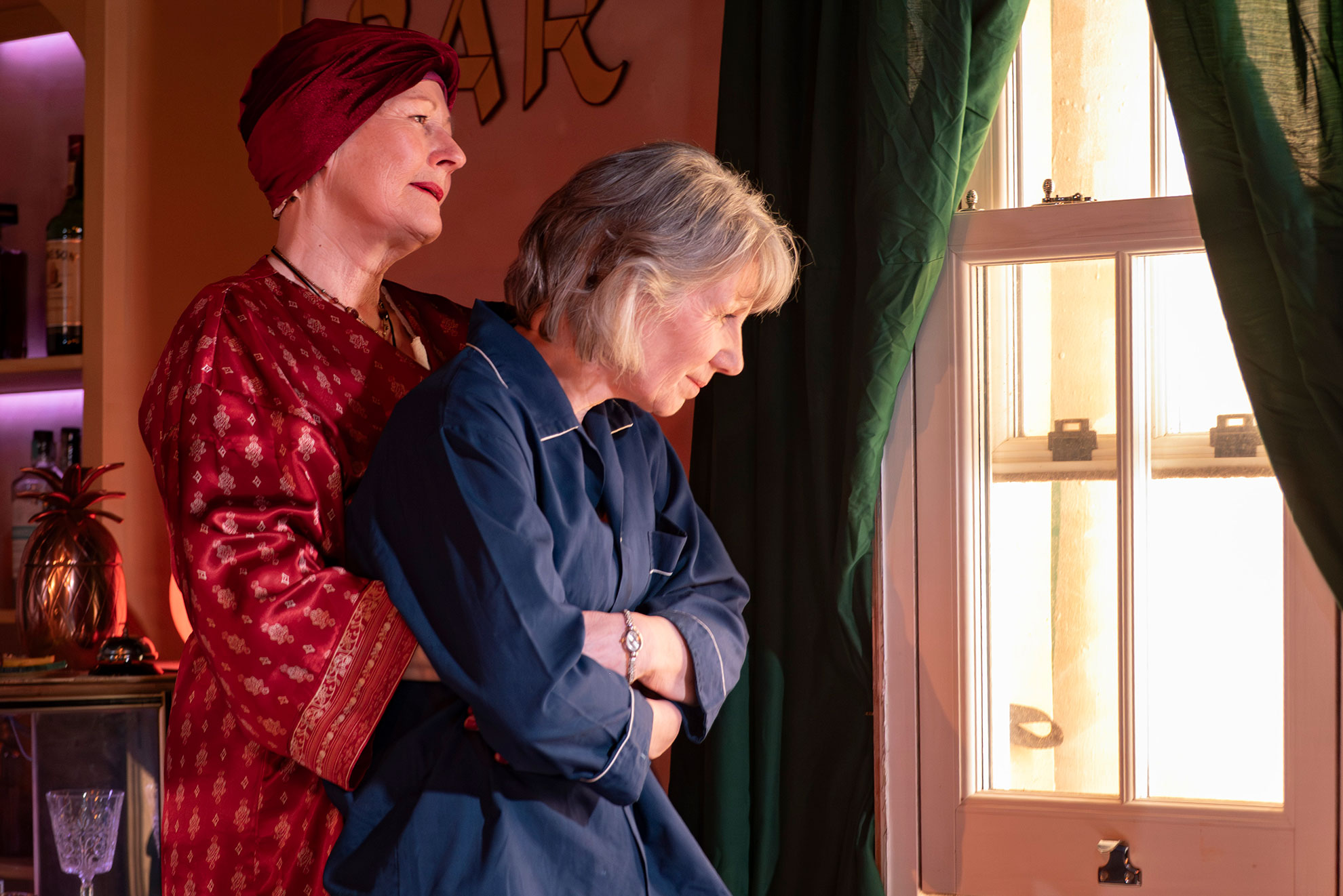 Two women stand by a window; the woman in red stands behind the grey-haired woman in blue, who looks out pensively.